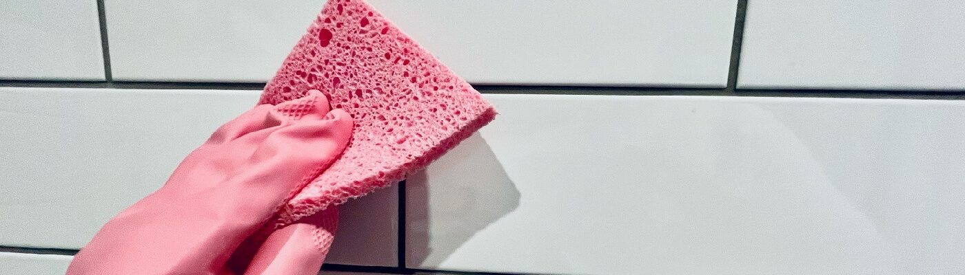A hand wearing a pink rubber glove holds a pink sponge is cleaning tiles (white metro tiles)