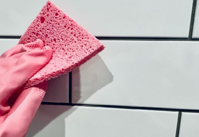 A hand wearing a pink rubber glove holds a pink sponge is cleaning tiles (white metro tiles)