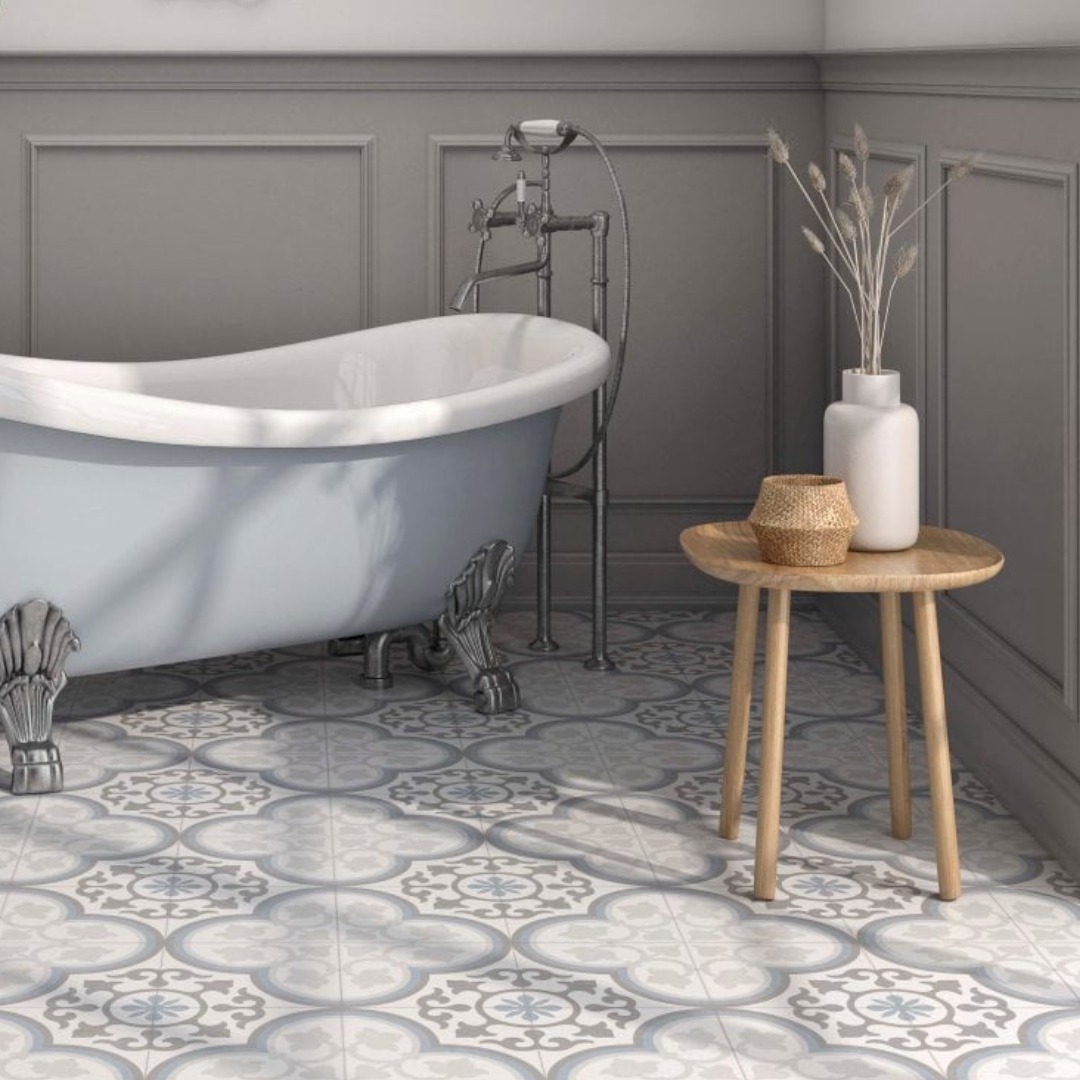 Close up of a bathroom with a large grey bath tub on the left and chelsea victorian patterned floor tiles. A small light brown table is on the right with a vase.