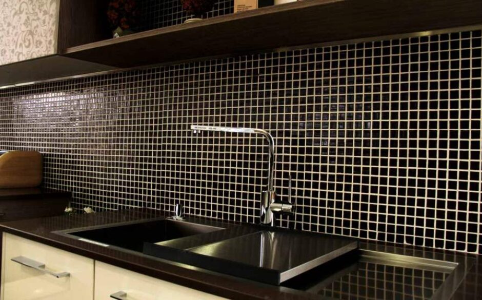 Image showing Black Mosaic Tiles on a wall in a kitchen with a large silver tap in front of the wall.