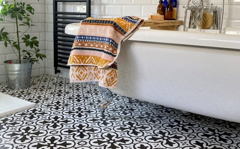 Photo of a bathtub with an Aztec style bath towel hanging over the edge of the bath. The focus of the photo is on the Boulevard Black and White Patterned Floor Tiles which have a vintage look. A black towel rack sits to the left of the bath in the backdrop of the photo against white metro style tiles.