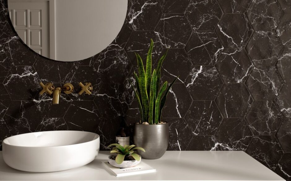 Photo of a black marble wall with hexagon shaped Dorset Black Hexagon Marble Tiles. A plant sits on a sink tabletop with a large round mirror in the top left corner of the wall. Brass taps come out of the wall hanging over a white dome shaped sink.